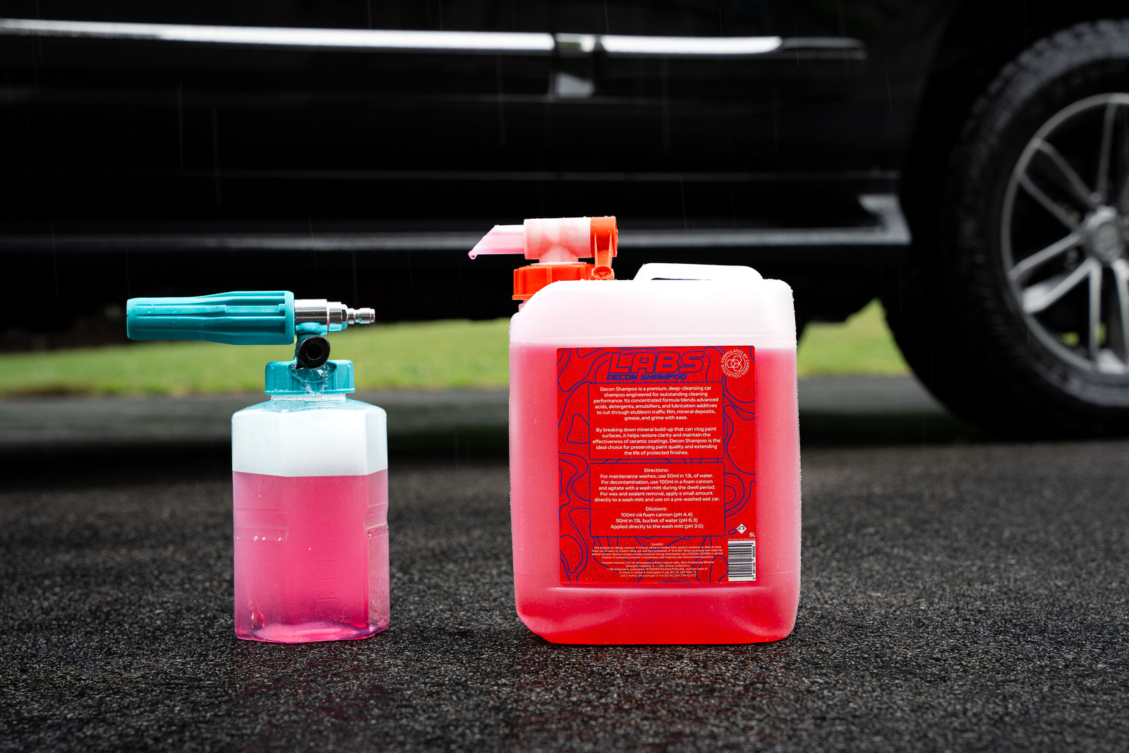LABS Decon Shampoo A diluted pink cleaning foam bottle with a turquoise foam cannon attachment and a large white plastic canister filled with diluted pink liquid, placed on asphalt in front of a blurred black vehicle, 5L jug