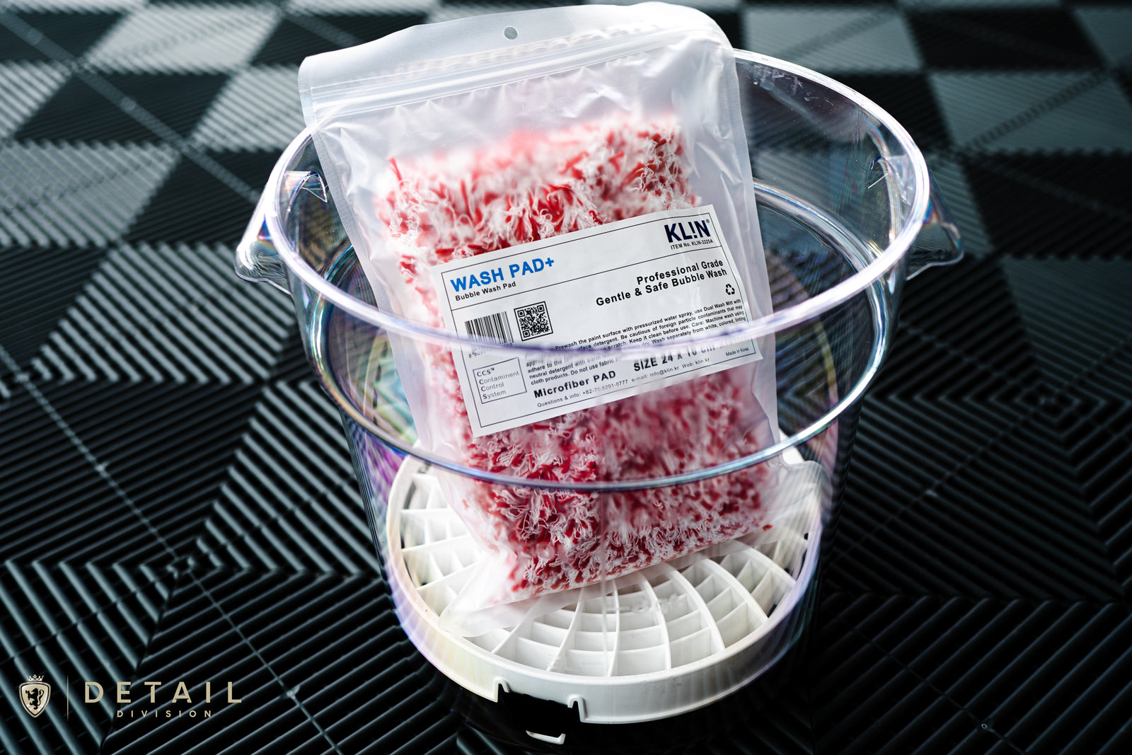 A clear, durable GHÖST brand wash bucket made of polycarbonate, shown with a white insert containing red and white wash pad, placed on a black textured swisstrax floor tiles.